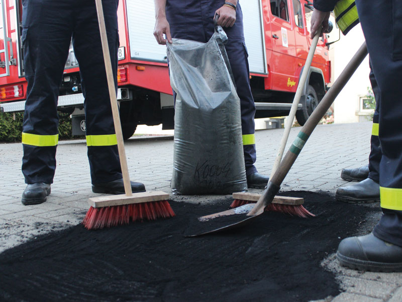sagu® additiv MYGRAN Vier Feuerwehrleute mit Besen und Schaufel nehmen mit Mygran-Ölbindemittel Öl von einer Fläche auf